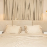 Lit avec tête de lit beige tapissée entre deux tables de chevet blanches, pour illustrer quelle taille de tête de lit choisir