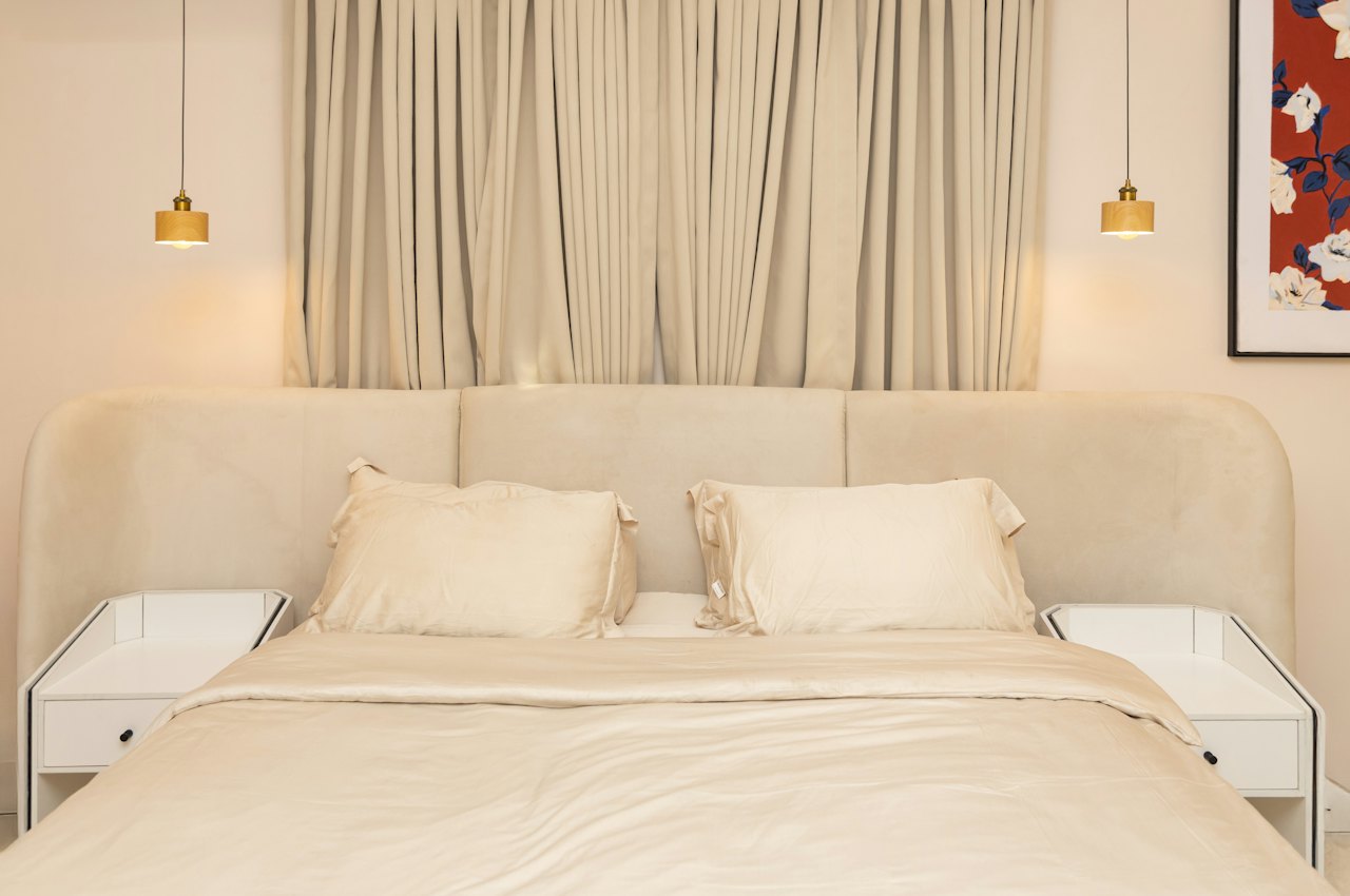 Lit avec tête de lit beige tapissée entre deux tables de chevet blanches, pour illustrer quelle taille de tête de lit choisir